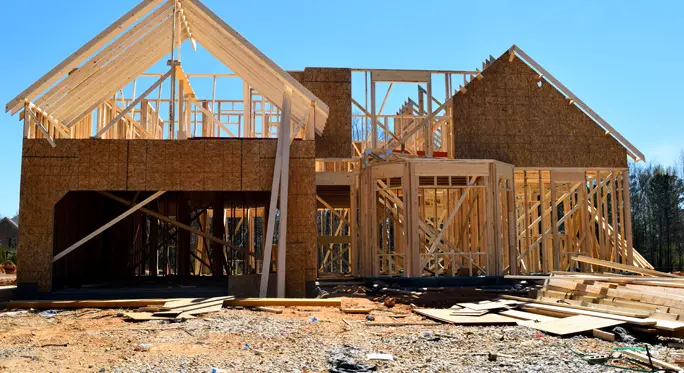 A large house under construction with exposed wooden framing and partially completed exterior walls on a clear day. Building materials and debris are scattered around the site.